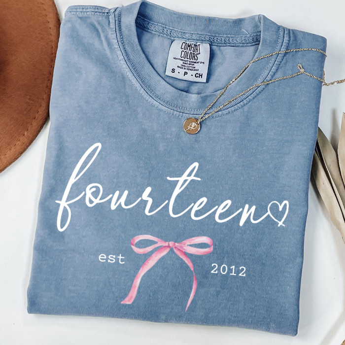 A folded light blue comfort colors t-shirt with a custom white 'fourteen' script, a pink ribbon graphic, and 'est 2012' printed on the front. A gold necklace is draped over the collar, and the shirt is styled with a brown hat on a white surface.