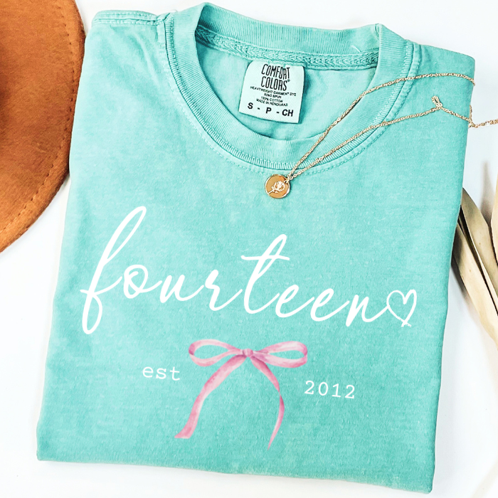 A folded mint green comfort colors t-shirt with 'fourteen' and 'est 2012' printed in white, decorated with a pink bow graphic. A gold pendant necklace is draped over the collar.