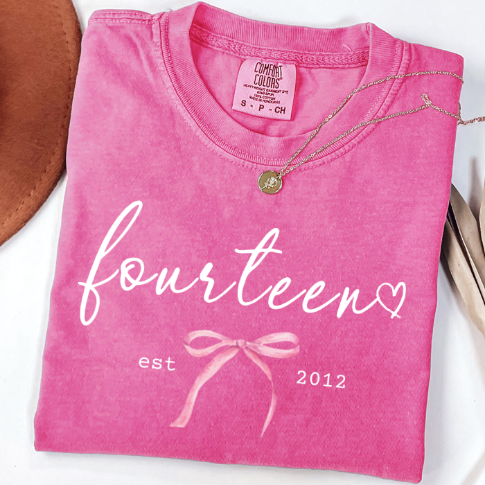 A folded bright pink comfort colors t-shirt with 'fourteen est 2012' and a bow graphic printed in white and light pink on the front. A gold necklace is draped over the collar, and a brown leather item is partially visible on the left.