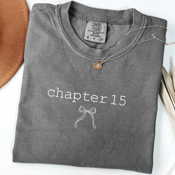 A folded charcoal gray comfort colors t-shirt with 'chapter 15' and a bow graphic printed in white on the front. A gold Bitcoin necklace is draped over the collar.