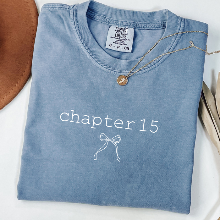 A folded light blue comfort colors t-shirt with 'chapter twelve' and five heart outlines printed on the front, styled with a delicate gold necklace on a white background.