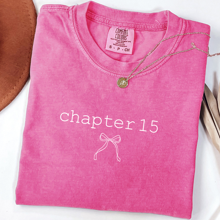 A folded bright pink comfort colors t-shirt with 'chapter 15' and a bow graphic printed in white on the front. A gold necklace is draped over the collar, and the shirt is styled with a brown leather item on a white surface.