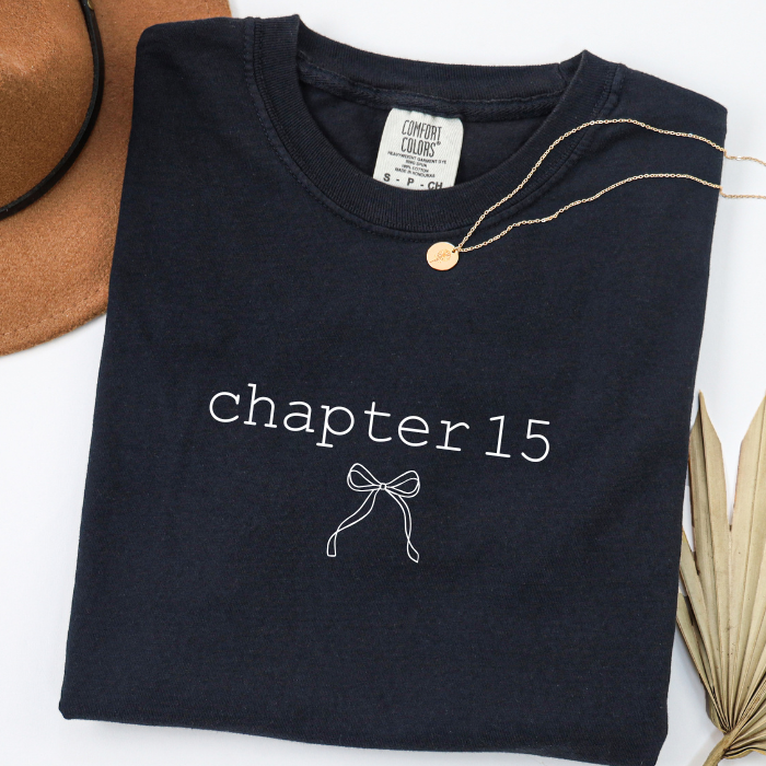 A folded black comfort colors t-shirt with 'chapter 15' and a bow graphic printed in white on the front. A gold necklace is draped over the collar, and a brown hat and dried palm leaf are styled as props on a white background.