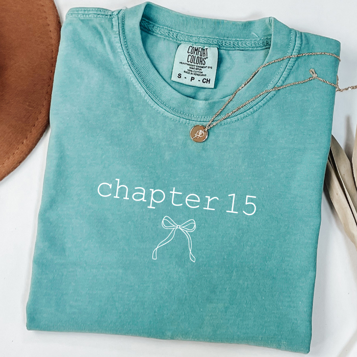 A folded turquoise crewneck comfort colors t-shirt with 'chapter 15' and a bow graphic printed on the front in white. A gold pendant necklace is draped over the collar.