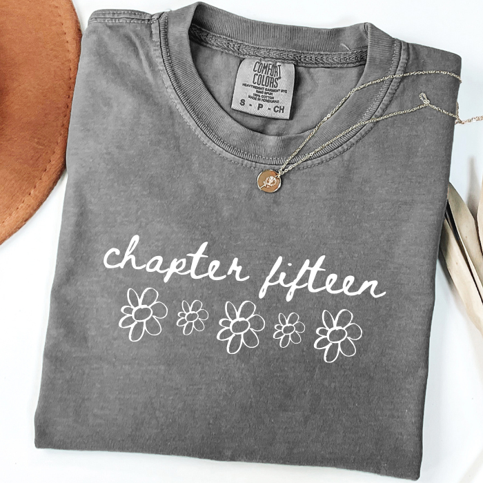 A folded gray comfort colors t-shirt with 'chapter fifteen' and flower graphics printed in white on the front, styled with a small gold pendant necklace on a white background.