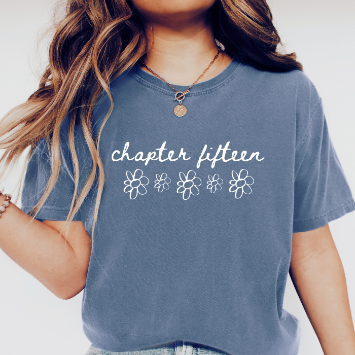 A person wearing a faded slate blue graphic comfort colors t-shirt with the words 'chapter fifteen' and five white outline flowers printed on the front.