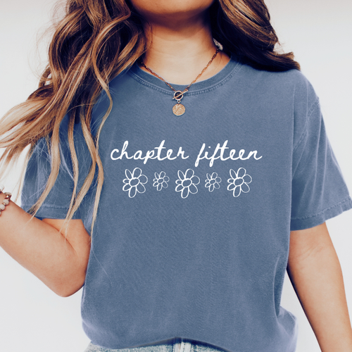 A person wearing a faded slate blue graphic comfort colors t-shirt with the words 'chapter fifteen' and five white outline flowers printed on the front.