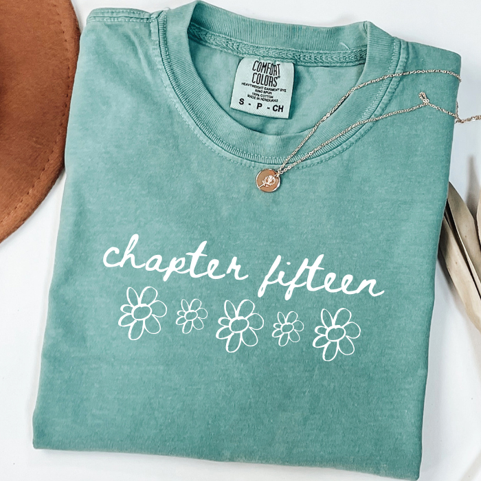 A folded mint green comfort colors t-shirt with 'chapter fifteen' and white flower graphics printed on the front, styled with a necklace on a white background.