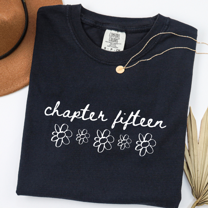 A folded black comfort colors t-shirt with 'chapter fifteen' and five white outlined flowers printed on the front, styled with a gold necklace, a brown hat, and dried palm leaves on a white background.