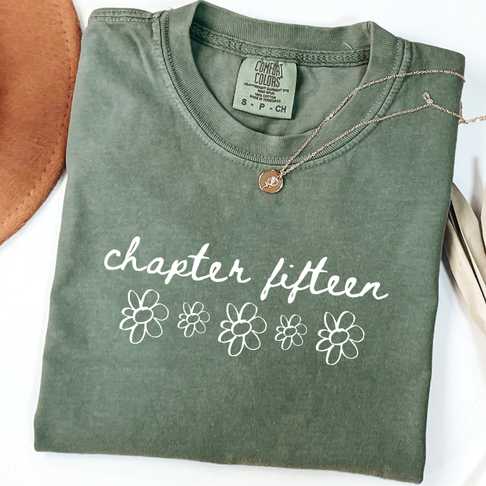 An olive green folded comfort colors t-shirt with 'chapter fifteen' and flower graphics printed on the front, styled with a necklace on a white background.
