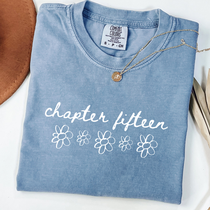 A folded light blue comfort colors t-shirt with 'chapter ten' and five heart outlines printed in white on the front. A delicate gold necklace is draped over the collar.