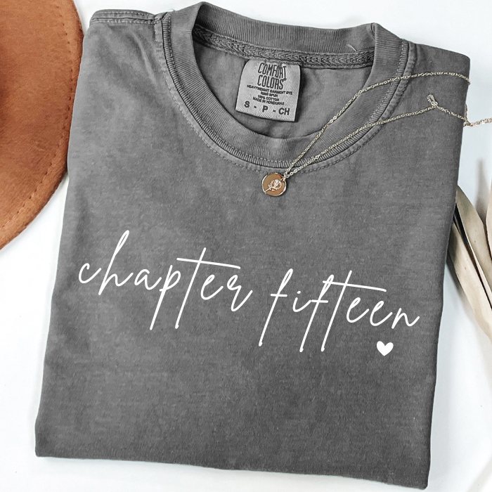 A folded dark gray comfort colors t-shirt with 'chapter fourteen' printed in white script across the front, styled with a small bronze pendant necklace on a white background with decorative props.
