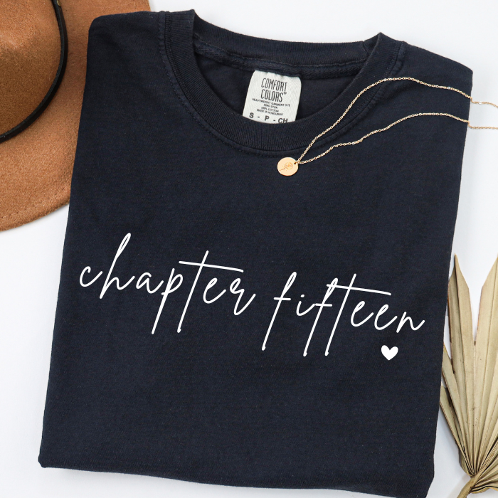 A folded black comfort colors t-shirt with 'chapter fifteen' printed in white script across the front, styled with a gold necklace on a white background with decorative props.