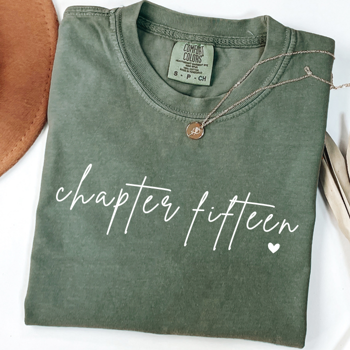 An olive green folded comfort colors t-shirt with 'chapter fifteen' printed in white script across the front, styled with a delicate necklace and surrounded by neutral props.