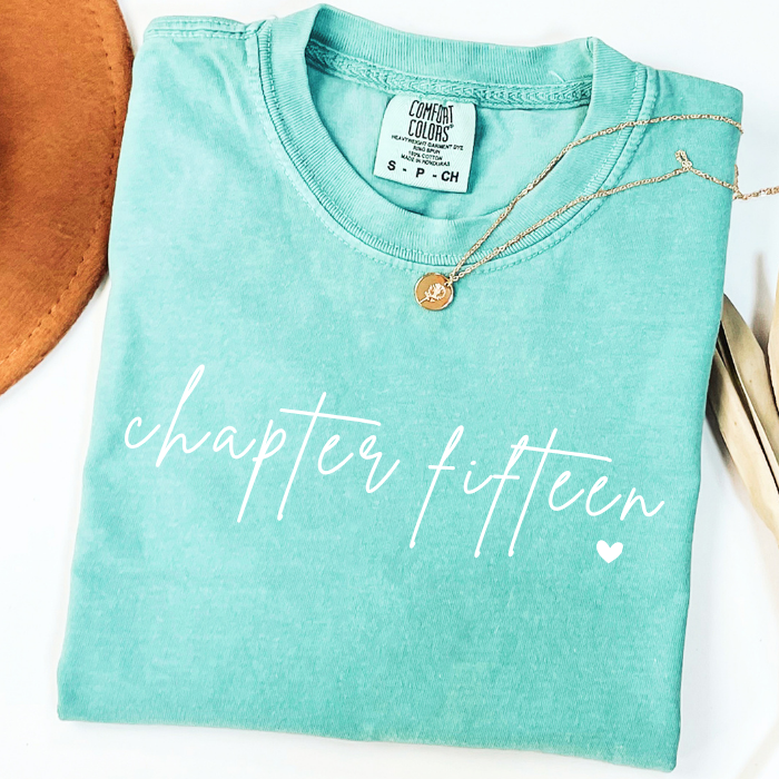 A folded mint green comfort colors t-shirt with 'chapter ten' printed in white script across the front, styled with a gold necklace and a brown leather accessory on a white background.