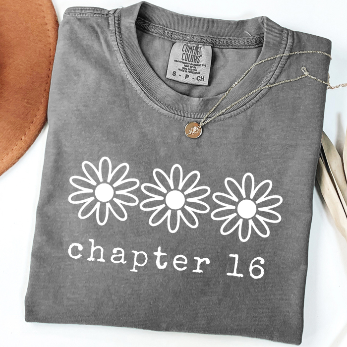 A folded dark gray comfort colors t-shirt with a white graphic print of three daisies and the text 'chapter 16' on the front. A small round pendant necklace is draped over the collar.