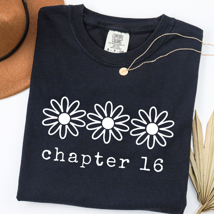 A folded black comfort colors t-shirt with three white daisy graphics and the text 'chapter 16' printed on the front. A gold necklace is draped over the collar, with a brown hat and dried palm leaves partially visible in the background.