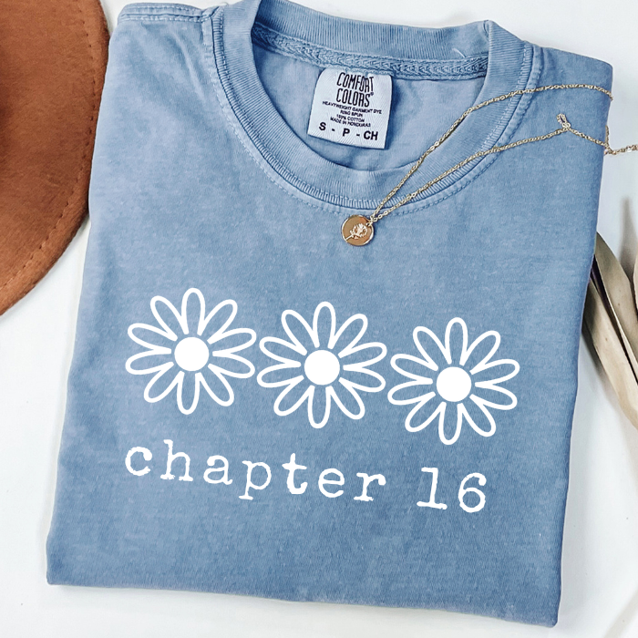 A folded light blue comfort colors t-shirt with three white daisy graphics and the text 'chapter 16' printed on the front. A gold necklace is draped over the collar, and a brown leather item is partially visible on the left side, all on a white background.