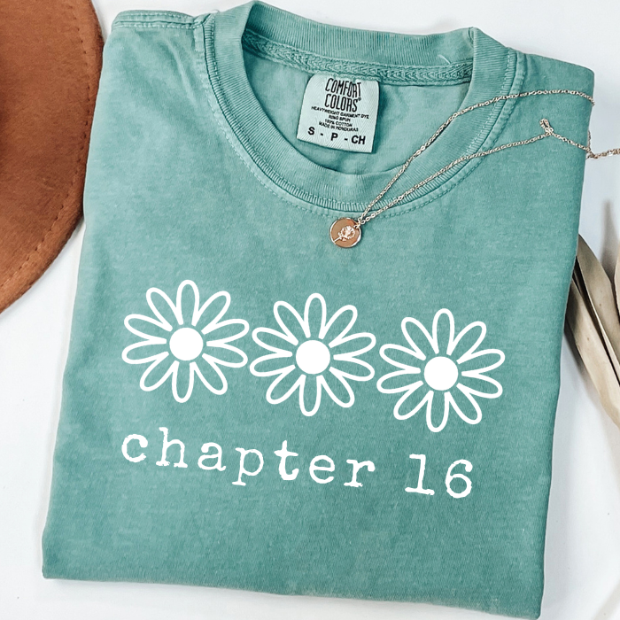 A folded mint green comfort colors t-shirt with a white graphic of three daisy flowers and the text 'chapter 16' printed on the front. A silver necklace with a Bitcoin pendant is draped over the collar.