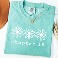 A folded mint green comfort colors t-shirt with a white graphic of three daisy flowers and the text 'chapter 16' printed on the front. A gold pendant necklace is draped over the collar.