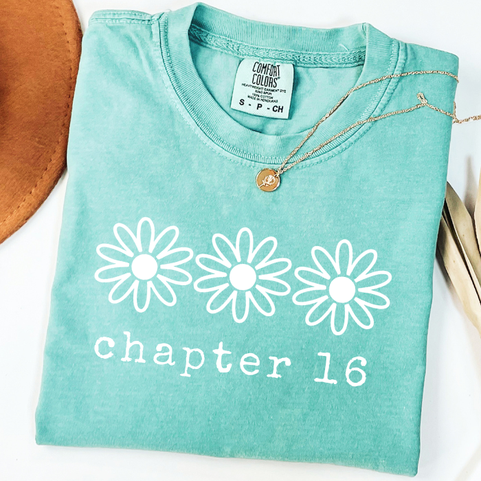 A folded mint green comfort colors t-shirt with a white graphic of three daisy flowers and the text 'chapter 16' printed on the front. A gold pendant necklace is draped over the collar.
