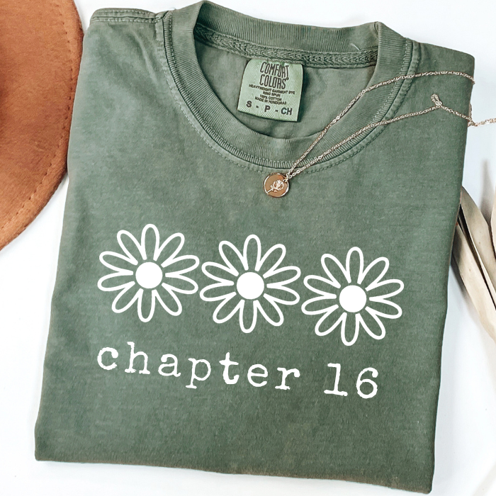 An olive green folded comfort colors t-shirt with a white graphic print of three daisy flowers and the text 'chapter 16' underneath. A gold necklace with a round pendant is draped over the collar.