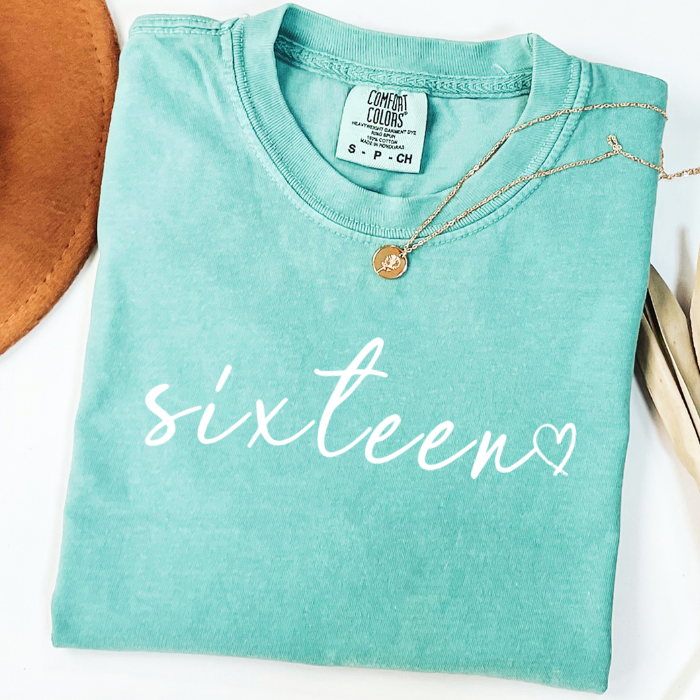 A folded mint green comfort colors t-shirt with 'sixteen' printed in white script across the front, styled with a gold necklace on a white background.