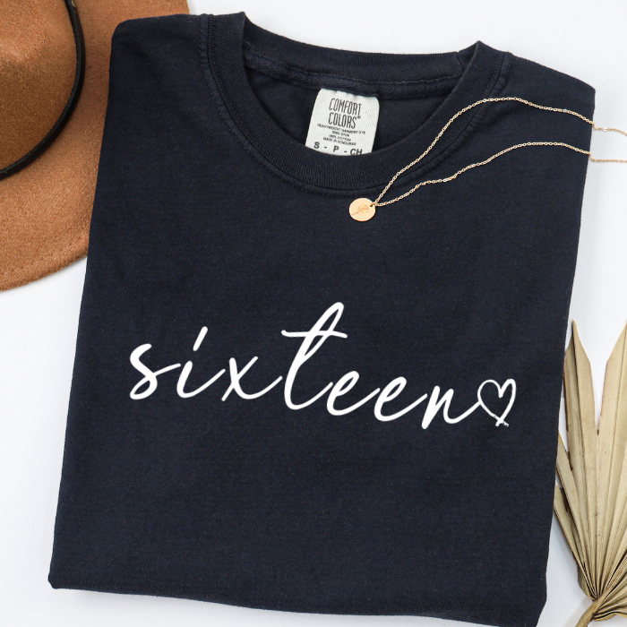 A folded black comfort colors t-shirt with 'sixteen' written in white script across the front, styled with a gold necklace on a white background with a brown hat and dried palm leaves.