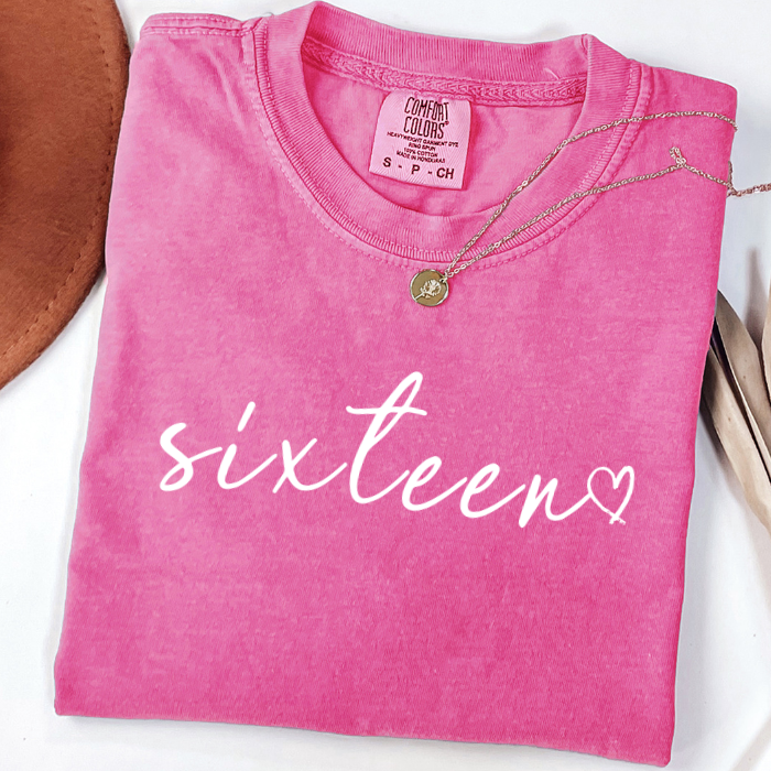 A folded bright pink comfort colors t-shirt with 'sixteen' printed in white script across the front, styled with a delicate gold necklace on a white background.
