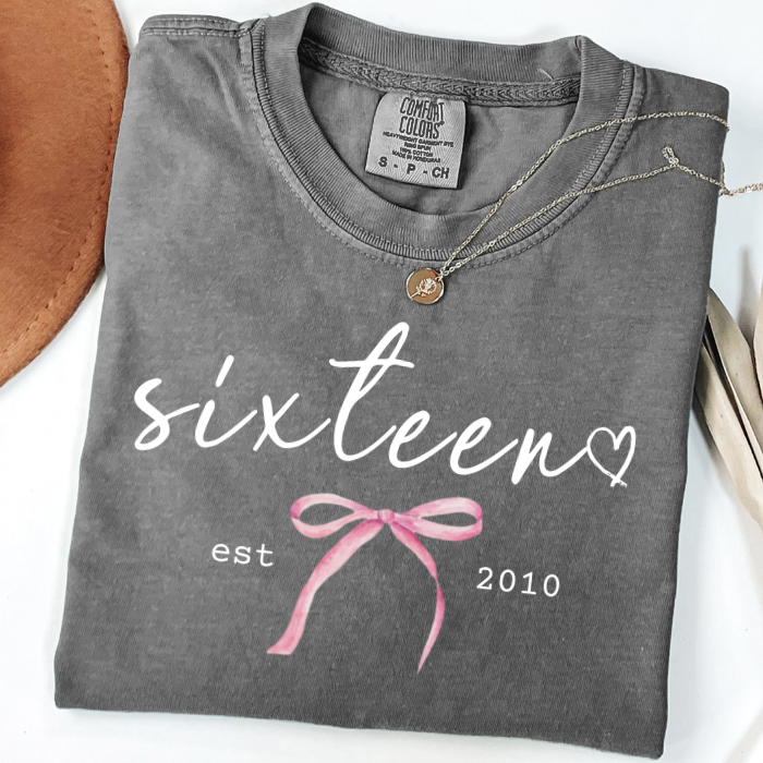 A folded dark gray graphic comfort colors T-shirt with white script text reading 'sixteen' and 'est 2010', plus a pink bow illustration. A small round pendant necklace is draped over the collar.