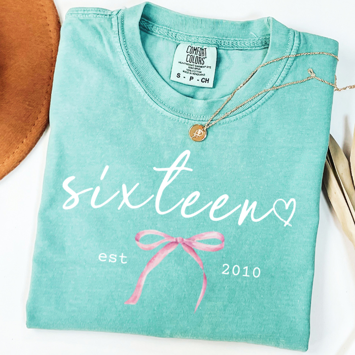A folded mint green comfort colors t-shirt with a graphic print that reads 'sixteen est 2010' and a pink bow illustration. A gold pendant necklace is draped over the collar.