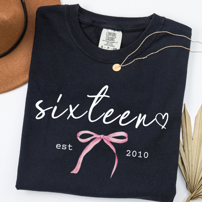A folded black comfort colors t-shirt with a white 'sixteen' script, a pink ribbon graphic, and 'est 2010' printed on the front. A gold necklace is draped over the collar, with a brown hat and dried palm leaves partially visible in the background.