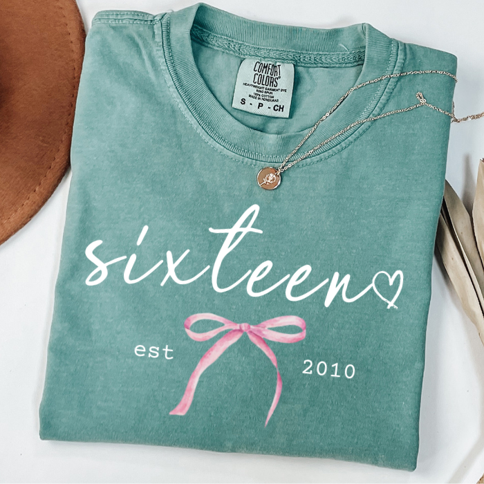 A folded mint green comfort colors t-shirt with 'sixteen est 2010' and a pink bow graphic printed on the front, styled with a necklace and props on a white surface.