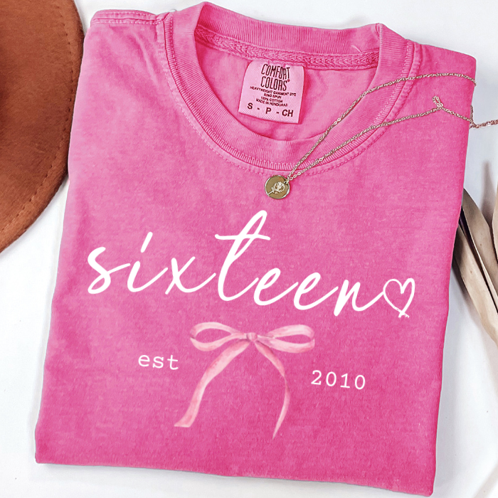 A folded bright pink comfort colors t-shirt with a mineral wash finish, featuring a white graphic print that reads 'sixteen' in script with a heart, 'est 2010' on either side, and a pink ribbon bow illustration. A gold necklace is draped over the collar.