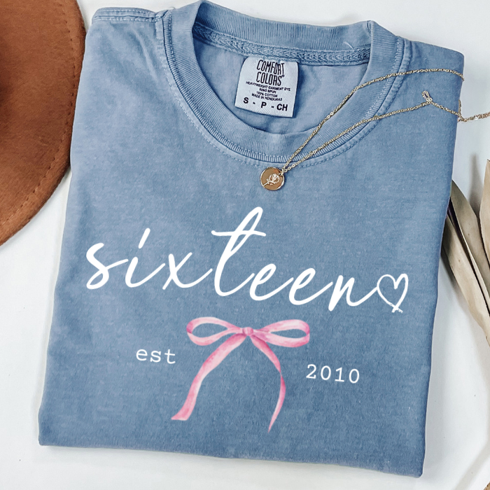 A folded light blue comfort colors t-shirt with 'sixteen' and 'est 2010' printed in white, decorated with a pink bow graphic. A gold necklace is draped over the collar, and a brown leather hat edge is visible on the left.