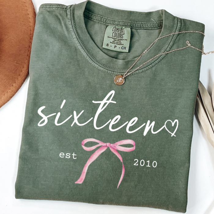 A folded olive green comfort colors t-shirt with a graphic that reads 'sixteen est 2010' in white, decorated with a pink ribbon illustration and a small heart. A delicate gold necklace is draped over the collar.