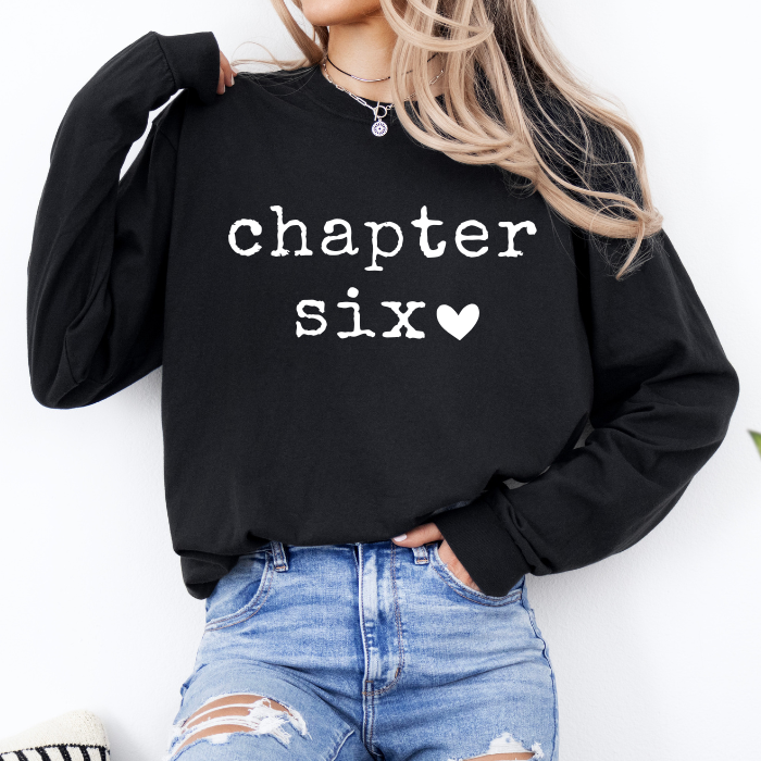 A woman wearing a black long-sleeve sweatshirt with 'chapter six' printed in white text and a small white heart, paired with distressed blue jeans.