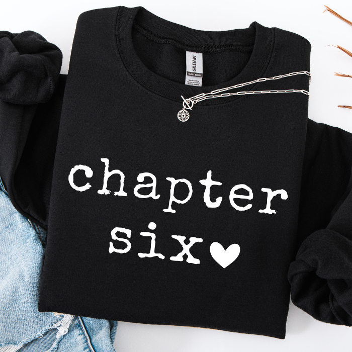 A black crewneck sweatshirt with 'chapter six' printed in white typewriter-style text and a white heart, styled flat-lay with a silver chain necklace and light blue denim.