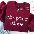 A folded burgundy sweatshirt with 'chapter six' printed in white across the front, styled with a silver chain necklace and light blue denim on a white background.