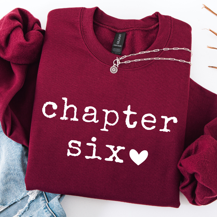 A folded burgundy sweatshirt with 'chapter six' printed in white across the front, styled with a silver chain necklace and light blue denim on a white background.
