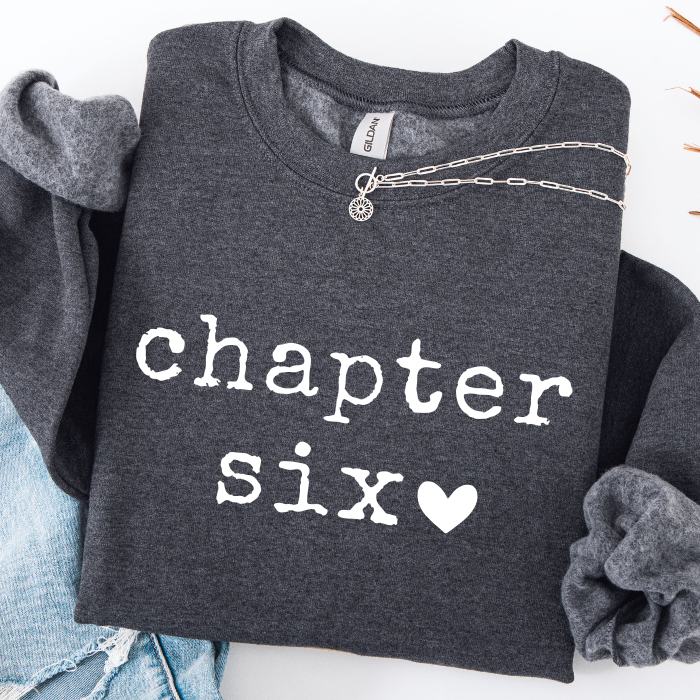 A dark gray sweatshirt with 'chapter six' printed in white typewriter-style text, styled flat-lay with a silver chain necklace, blue jeans, and a gray scrunchie on a white background.