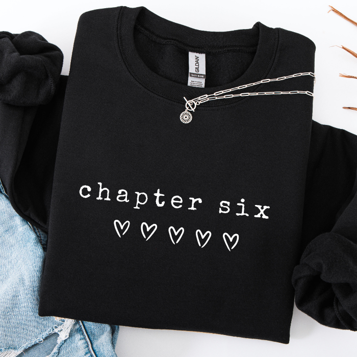 A folded black crewneck sweatshirt with 'chapter six' and five heart icons printed in white on the front. A silver chain necklace is draped across the chest.