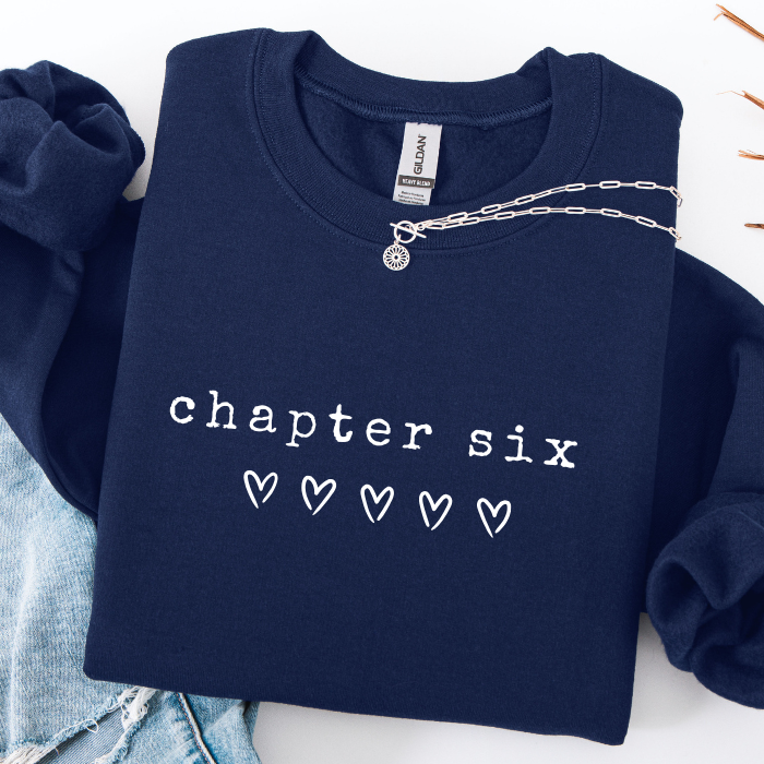 A navy blue sweatshirt with 'chapter six' and five outlined hearts printed in white across the chest, styled flat on a white surface with a silver chain necklace draped across the collar and light blue denim partially visible at the side.