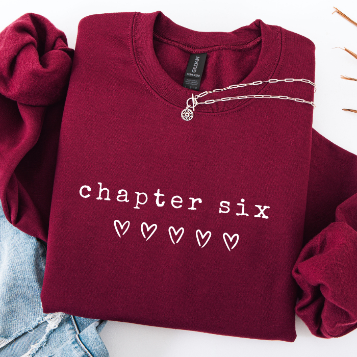 A folded burgundy sweatshirt with 'chapter six' and five heart graphics printed on the front, styled with a silver chain necklace and light blue denim on a white background.