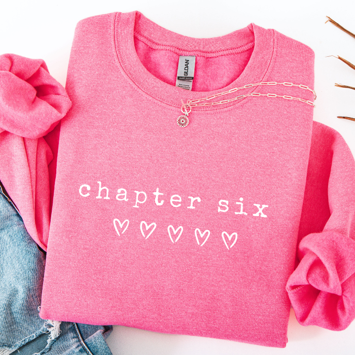A folded bright pink crewneck sweatshirt with 'chapter six' and five outlined hearts printed on the front, styled with rolled sleeves, a gold chain necklace, and a denim jacket on a white background.