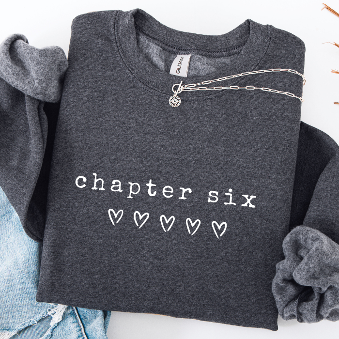 A dark heather gray sweatshirt with 'chapter six' and five hearts printed in white on the front, styled flat-lay with rolled sleeves, a silver chain necklace, and light blue jeans on a white background.