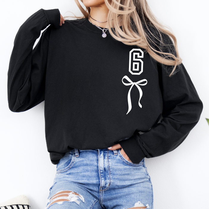 A woman wearing a black long-sleeve sweatshirt with a white outlined number 6 and bow graphic on the front, paired with distressed blue jeans.