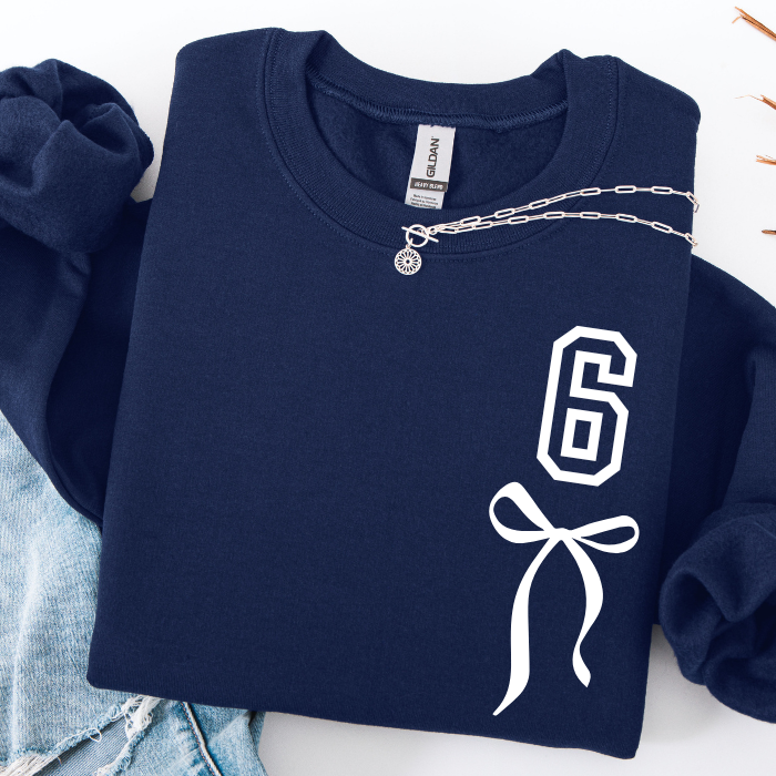 A navy blue sweatshirt with a white graphic print of the number 6 and a bow on the front, styled flat-lay with a silver chain necklace and light blue denim jeans.