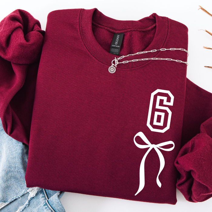 A folded burgundy crewneck sweatshirt with a white graphic print of the number 6 and a ribbon bow on the front. A silver chain necklace with a small charm is draped across the collar area.