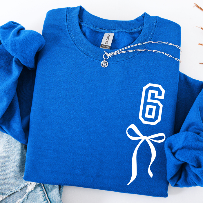 A folded royal blue sweatshirt with rolled sleeves, featuring a white outlined number 6 and bow graphic on the front. A silver chain necklace is draped across the chest, and light-wash denim jeans are partially visible underneath on a white background.